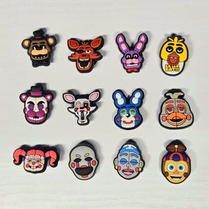 May include: A collection of colorful cartoon character charms, featuring various animal and humanoid faces. The charms are in shades of brown, red, purple, yellow, and blue, with black and white accents. Each charm has a unique design and expression.