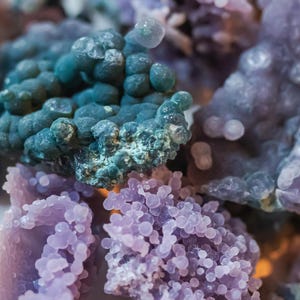May include: Close-up of a cluster of purple, blue, and green agate stones. The stones have a rough, textured surface and are arranged in a random pattern.