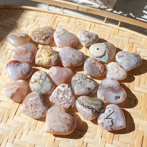 Puede incluir: Una colección de 17 piedras de ágata en forma de corazón en varios tonos de rosa, blanco y marrón con intrincados patrones. Las piedras están dispuestas en una bandeja de bambú tejido.