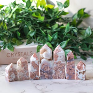 Puede incluir: Siete puntas de cristal de ágata rosa y blanca dispuestas en fila sobre una superficie de mármol blanco. Los cristales son de diferentes tamaños y formas, y tienen un aspecto natural y orgánico. La palabra "CRYSTALS" es visible en una caja detrás de los cristales.