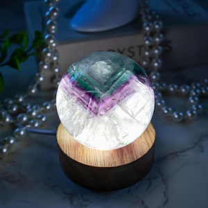 May include: A glowing crystal ball with green, purple, and white hues sits atop a wooden base. The sphere is illuminated from within, creating a soft, ambient light. Decorative pearls and a plant are in the background.
