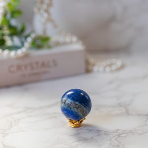 May include: A blue lapis lazuli sphere on a gold stand. The sphere is a deep blue with white and grey veins. The stand is a simple design with a round top and three legs.