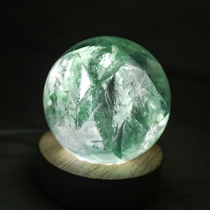 May include: A glowing, spherical crystal sculpture with a rough, textured surface. The crystal is primarily green with white and clear areas, mounted on a round wooden base. The light source is internal, creating a soft glow.