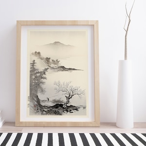 Puede incluir: Una pintura de tinta negra y blanca de un paisaje con montañas, árboles y una figura pescando en un lago. La pintura está enmarcada en un marco de madera clara y está colgada en una pared blanca.