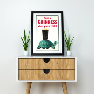 Op de afbeelding: Een zwart ingelijste poster met een vintage Guinness-reclame. De poster toont een groene schildpad die een glas Guinness draagt met de tekst "Have a GUINNESS when you're TIRED".