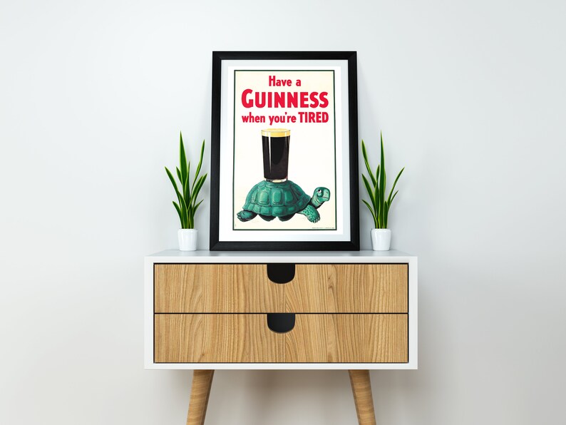 Op de afbeelding: Een zwart ingelijste poster met een vintage Guinness-reclame. De poster toont een groene schildpad die een glas Guinness draagt met de tekst "Have a GUINNESS when you're TIRED".