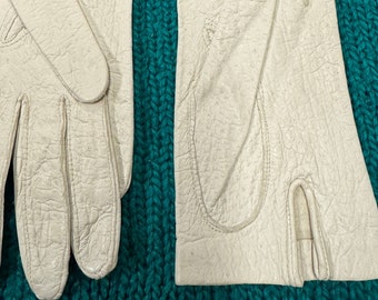 May include: A pair of white leather gloves with a stitched design. The gloves are laid flat on a green knitted surface.