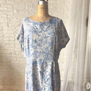 May include: A vintage blue dress with a white floral pattern. The dress has short sleeves, a fitted waist, and a knee-length skirt. It is displayed on a dress form against a white brick wall and dark wood floor.