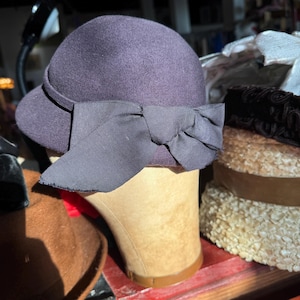 May include: A dark purple felt cloche hat featuring a matching dark purple grosgrain ribbon band that wraps around the crown and terminates in a large, structured bow detail on the side. The hat has a rounded crown and a small, downturned brim.