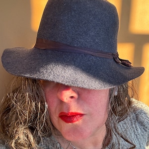 May include: A woman wearing a grey felt fedora with a brown ribbon. The hat is pulled down low, covering most of her face. She is wearing a grey sweater and has long, wavy hair.
