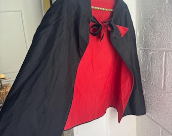 Könnte beinhalten: Ein Wendecape mit schwarzer Außenseite und roter Innenseite. Das Cape hängt an einem Kleiderbügel und ist am Hals mit einer Schleife gebunden. Es besteht aus einem leichten Stoff.