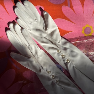 Peut inclure: Une paire de gants blancs, probablement pour des occasions formelles. Les gants sont faits d'un matériau lisse, probablement satiné. Chaque gant comporte trois boutons en forme de perle au poignet. Le fond est un motif floral coloré.