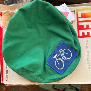 May include: A green cycling cap with a blue patch showing a white bicycle design. The cap is made of a solid green fabric. The cap is resting on a vintage magazine.