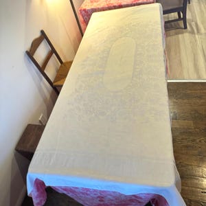 Op de afbeelding: Een grote, rechthoekige tafelkleed met een crèmekleurig bloemenpatroon. Het tafelkleed is over een tafel gedrapeerd, met een rood en wit patroonstof eronder zichtbaar. De tafel staat in een kamer met houten vloeren en een stoel.