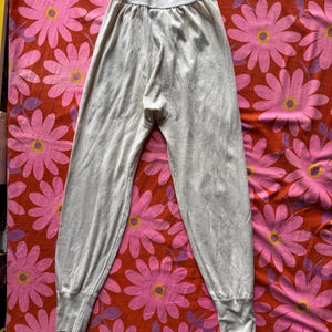 Könnte beinhalten: Eine hellbeige Thermohose mit geripptem Bund und Bündchen. Die Hose aus weichem Strickstoff wird auf einem floral gemusterten Hintergrund mit rosa und roten Blumen präsentiert.