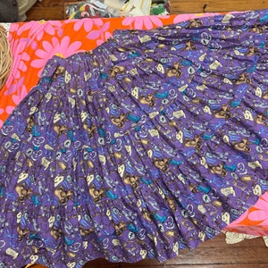 May include: A purple skirt with a pattern of cowboy boots, hats, and other western-themed imagery. The skirt is tiered and has a gathered waist.