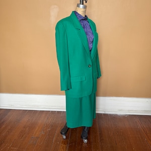 Puede incluir: Un traje de chaqueta y falda verde. La chaqueta tiene un cierre de botón único y un bolsillo a cada lado. La falda es de largo hasta la rodilla.