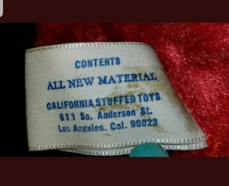 K&ouml;nnte beinhalten: Ein wei&szlig;es Etikett mit blauem Text, auf dem steht "CONTENTS ALL NEW MATERIAL CALIFORNIA STUFFED TOYS 611 So. Anderson St. Los Angeles. Cal. 90023".