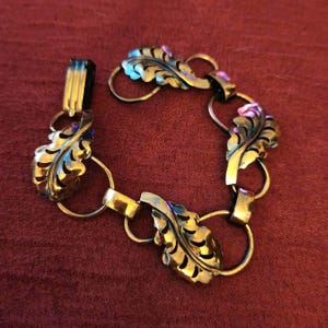 May include: A gold-toned bracelet featuring leaf-shaped links. The leaves have intricate vein detailing and are connected by circular and rectangular links. The clasp is a simple bar and loop design. The bracelet is displayed on a red surface.