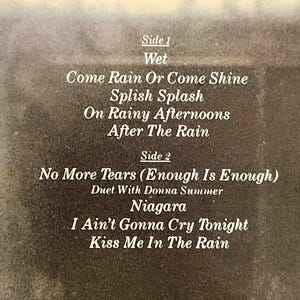 May include: A close-up of a vintage album cover with song titles in white text. The album is divided into Side 1 and Side 2, with song titles like "Come Rain Or Come Shine" and "No More Tears". The background is a faded brown.