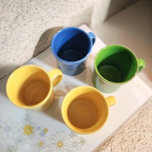 May include: Four colorful ceramic mugs in yellow, blue, and green are arranged on a white tray with a floral design. Each mug has a rounded shape with a handle. The mugs are empty and ready to be filled.