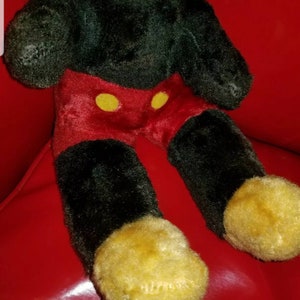 K&ouml;nnte beinhalten: Ein Pl&uuml;schtier Mickey Mouse in Schwarz und Rot, das auf einer roten Oberfl&auml;che sitzt. Das Spielzeug hat gelbe Kn&ouml;pfe auf seiner roten Hose und gelbe Schuhe.