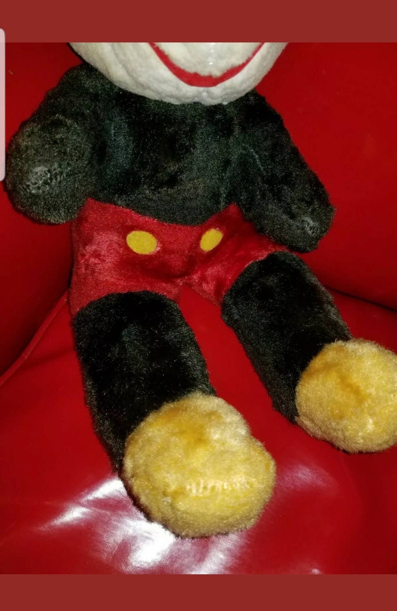 K&ouml;nnte beinhalten: Ein Pl&uuml;schtier Mickey Mouse in Schwarz und Rot, das auf einer roten Oberfl&auml;che sitzt. Das Spielzeug hat gelbe Kn&ouml;pfe auf seiner roten Hose und gelbe Schuhe.