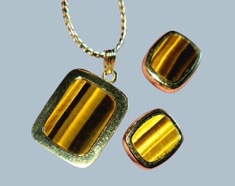 May include: A gold-toned jewelry set featuring a pendant necklace and matching earrings. The pendant and earrings have a rectangular and square shape, respectively, with a tiger's eye stone set in a gold frame. The necklace chain is also gold.