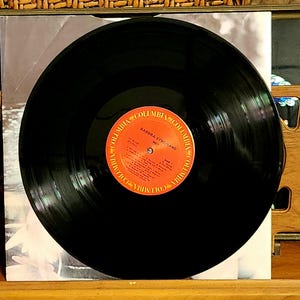 May include: A black vinyl record with a red and orange label featuring the text "Barbra Streisand Wet" and the Columbia Records logo. The record is centered on a white album cover, with a wooden surface in the foreground.