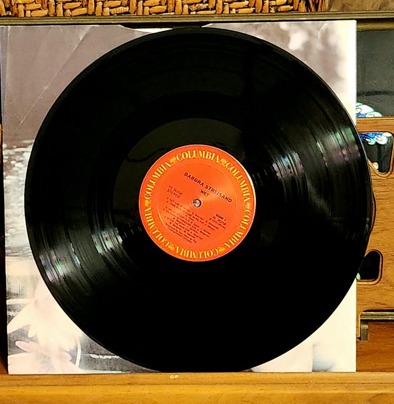 May include: A black vinyl record with a red and orange label featuring the text "Barbra Streisand Wet" and the Columbia Records logo. The record is centered on a white album cover, with a wooden surface in the foreground.
