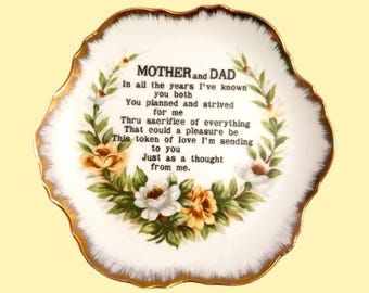 May include: A decorative white porcelain plate with a gold rim and floral accents. The plate features the text "MOTHER and DAD" and a poem. The flowers are white and yellow with green leaves.