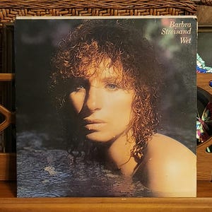 May include: Vintage vinyl record album cover featuring Barbra Streisand. The album cover has a dark background with Streisand's face and shoulders emerging from water. The album title "Wet" is visible.