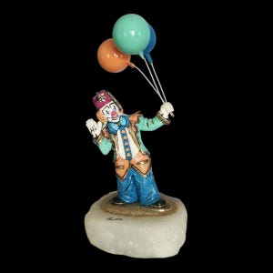May include: A painted clown figurine standing on a white stone base, wearing a purple hat, teal jacket, and blue trousers. The clown holds three balloons in orange, teal, and blue. The word Hi is written on the clown's white glove.