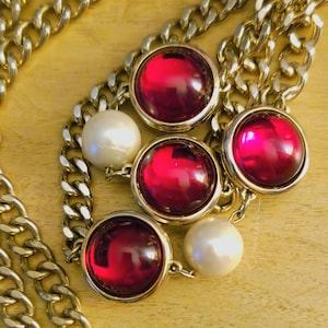 May include: A gold-tone chain necklace featuring large, round, ruby-red cabochons set in gold bezels. The necklace also includes large, round, white faux pearls. The chain is a chunky curb link style, creating a bold, statement piece.