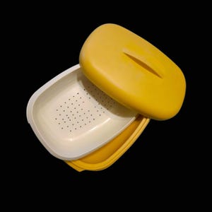 May include: A yellow and white plastic food storage container with a lid. The lid is yellow with a handle-shaped cutout. The container has a white interior with small holes. The container is open, revealing the interior.