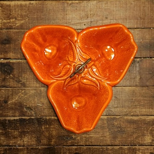 May include: Orange ceramic serving dish with three sections, shaped like a stylized strawberry. The dish has a textured surface with a central metal handle. The dish is set on a wooden surface.