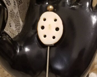 May include: A white oval-shaped pin with a gold top and five holes. The pin is on a black background.
