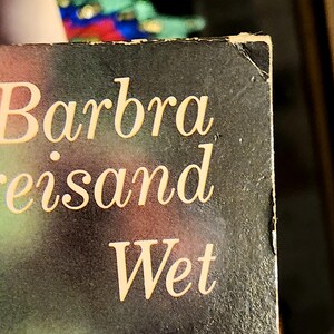 May include: Close-up of a book cover with the name "Barbra Streisand" and the title "Wet" in a gold, elegant font. The background is a dark, gradient color. The corner of the book shows some wear.