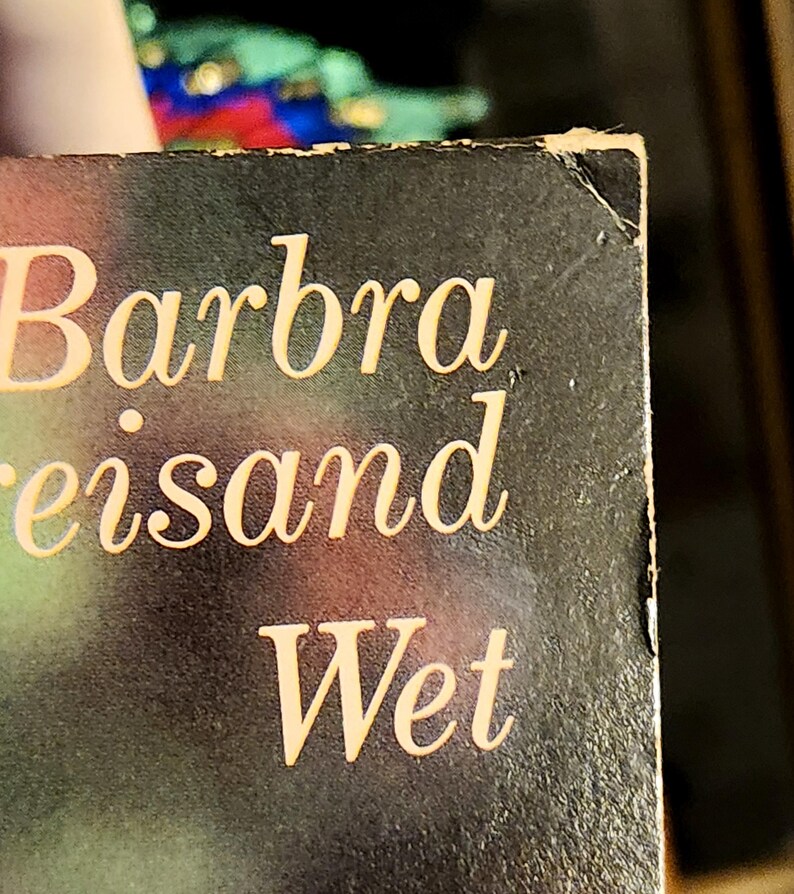 May include: Close-up of a book cover with the name "Barbra Streisand" and the title "Wet" in a gold, elegant font. The background is a dark, gradient color. The corner of the book shows some wear.
