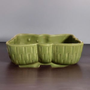 May include: A green ceramic planter with three connected compartments. The planter has a textured, vertical lined pattern on the exterior and a scalloped edge. The color is a muted, earthy green.