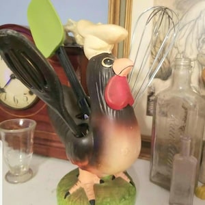 May include: A ceramic rooster kitchen utensil holder with a white chef's hat. The rooster is brown and black with a red comb and wattle. The holder has a green base and holds a green spatula, a black whisk, and a silver wire whisk.