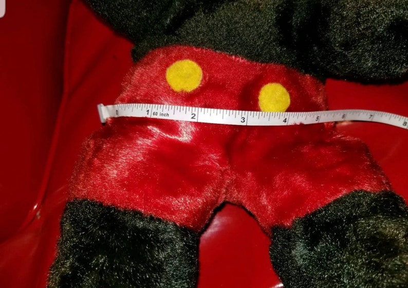 K&ouml;nnte beinhalten: Ein rotes und schwarzes Pl&uuml;sch-Mickey-Mouse-Spielzeug mit gelben Kn&ouml;pfen. Das Spielzeug wird mit einem Ma&szlig;band gemessen und zeigt, dass es 4 Zoll breit ist.