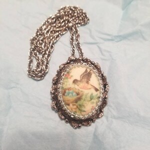 May include: A silver chain necklace with an oval pendant featuring a vintage-style image of a bird with a nest of eggs. The pendant is surrounded by a beaded border.