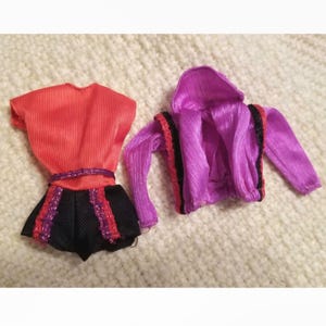 May include: A set of two doll clothes, one a red and black dress with a purple belt, and the other a purple jacket with black and red trim.