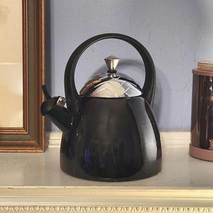 Puede incluir: Una tetera negra con una tapa plateada y un pomo en forma de diamante. La tetera tiene un acabado brillante y un pico curvo.