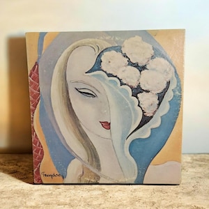May include: Square artwork featuring a stylized portrait of a woman with long blonde hair and red lips. The woman's face is partially obscured by a blue veil, and a cluster of white flowers is in the background. The artwork has a vintage aesthetic.