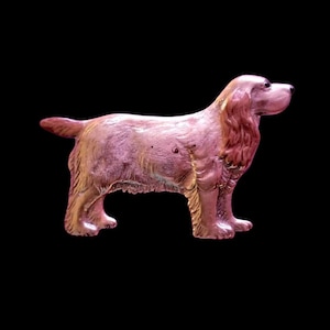 May include: A bronze-colored dog figurine, likely a Cocker Spaniel, stands in profile against a black background. The dog's fur is detailed, with a textured appearance. The figurine has a glossy finish, and the dog's tail is slightly curled.