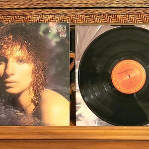 May include: Album cover for Barbra Streisand's "Wet" album. The cover features a portrait of Streisand in water, with the album title and artist name visible. A vinyl record is next to the album cover.