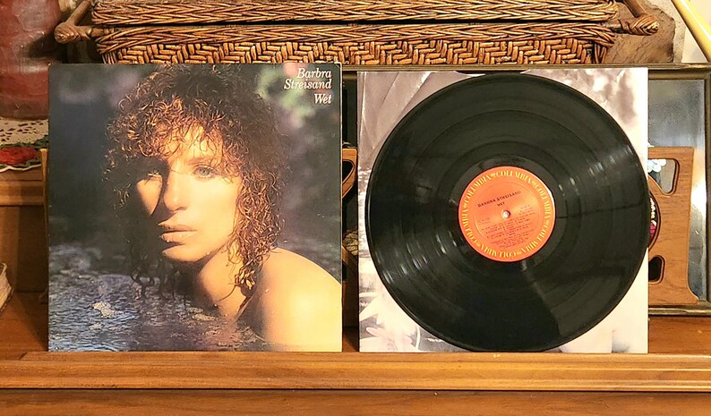May include: Album cover for Barbra Streisand's "Wet" album. The cover features a portrait of Streisand in water, with the album title and artist name visible. A vinyl record is next to the album cover.