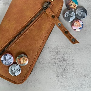 May include: A brown leather wallet with a zipper closure and a brown leather strap with metal studs. The wallet is decorated with several round pinback buttons featuring images of a woman with blonde hair.